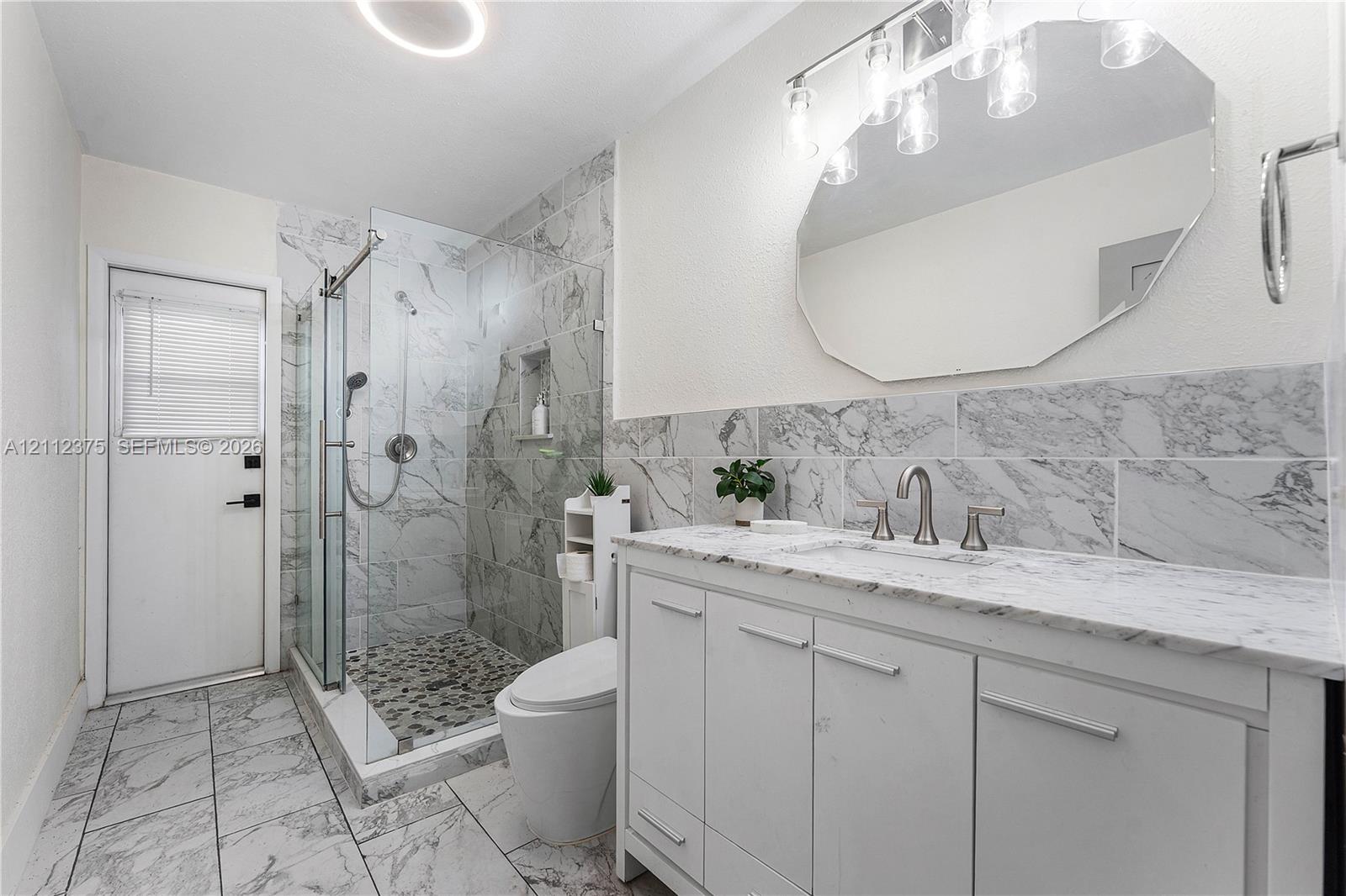 Bathroom featuring a white vanity with a stone countertop, an expansive walk-in shower with stone tile surround and pebble flooring, and a large octagonal wall mirror