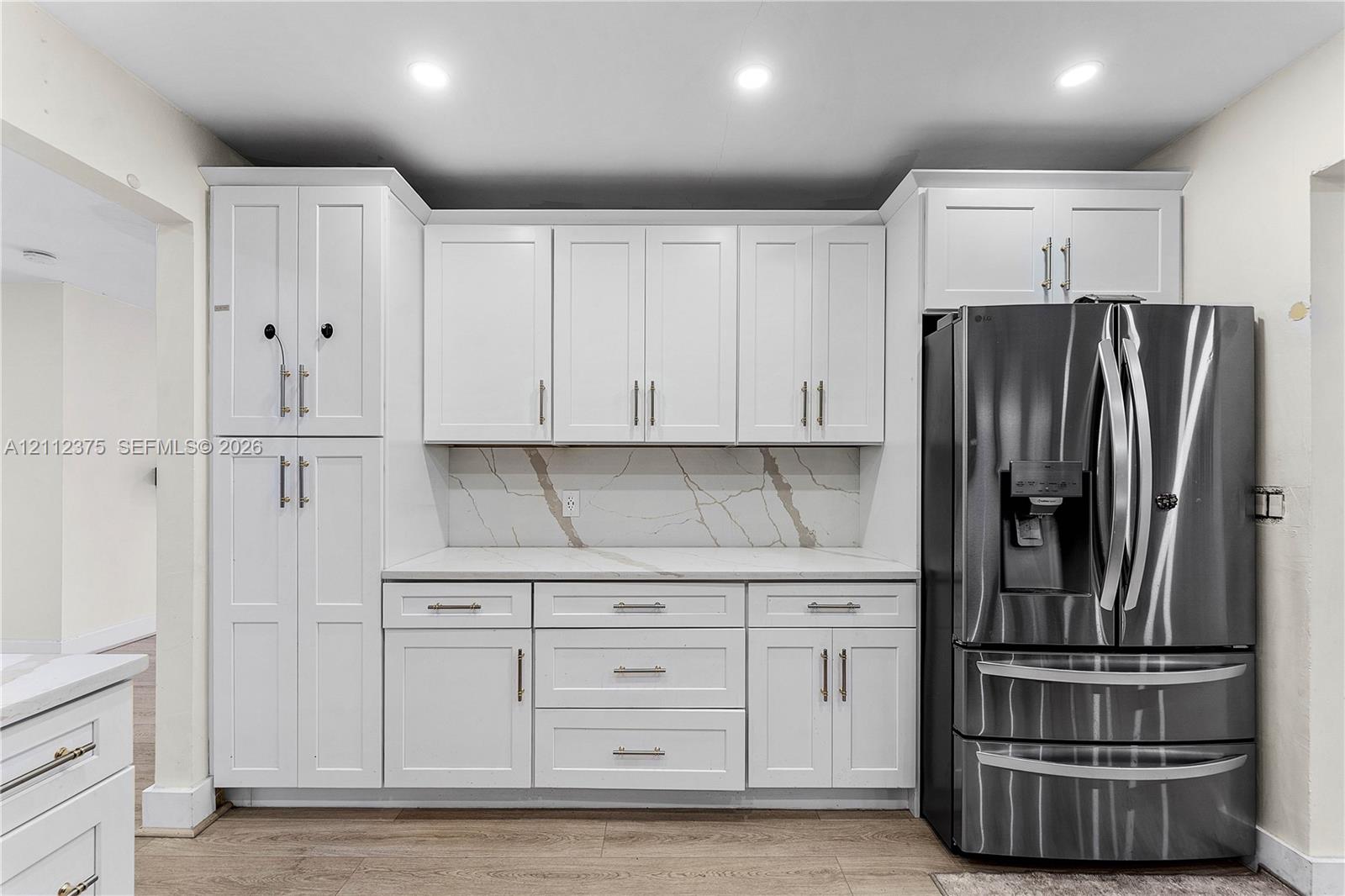 White shaker-style cabinetry with matte black hardware, stone-patterned backsplash, quartz countertops, stainless steel French door refrigerator, and wood-finish flooring
