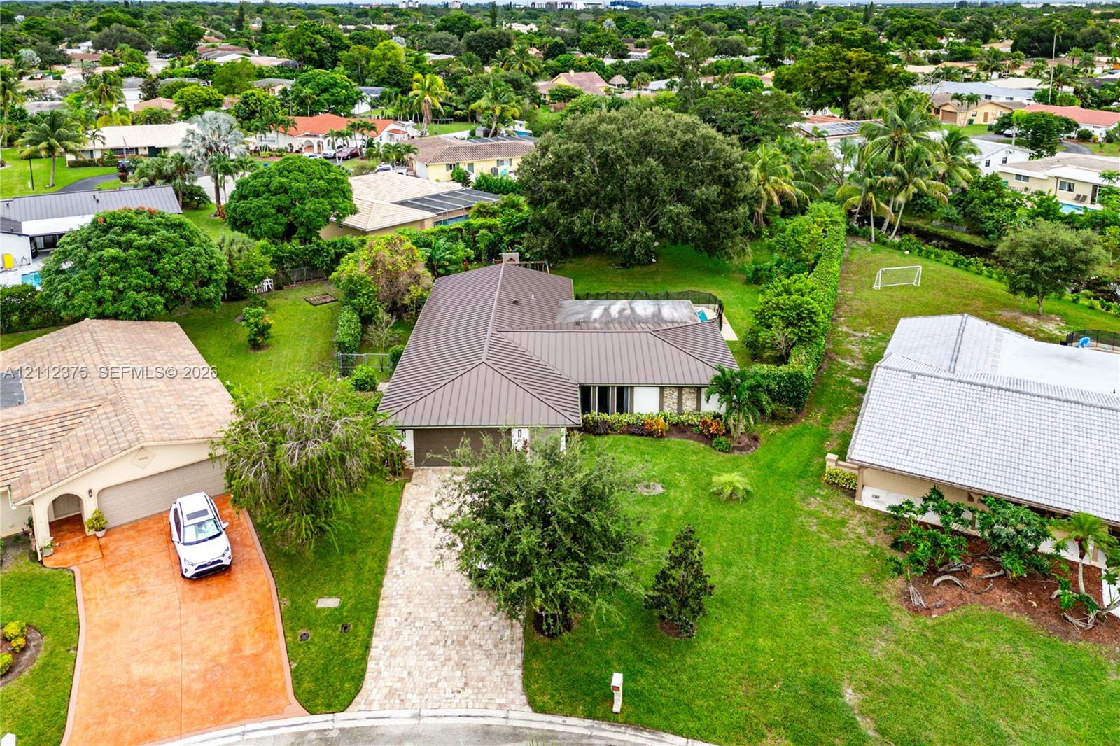 Aerial perspective featuring a metal roof, stone-paved driveway, and expansive lawn with mature landscaping