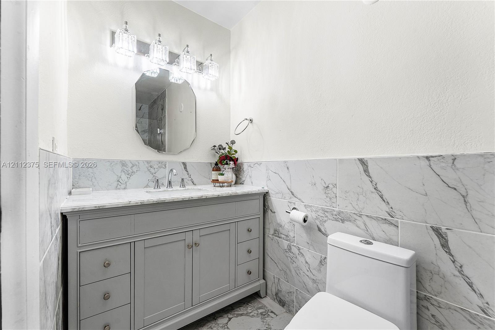 Grey vanity with stone-surface countertop, porcelain wall tile with integrated veining, octagonal wall mirror, and four-bulb polished vanity lighting