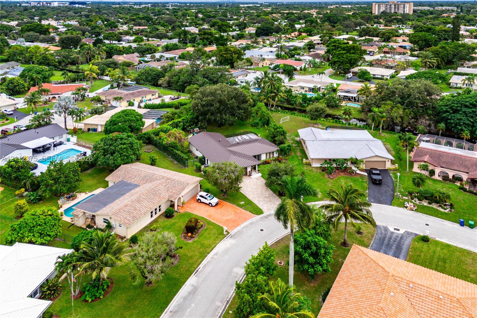 Aerial perspective of a suburban neighborhood featuring paved cul-de-sac streets, mature tropical landscaping, residential properties with shingled roofing, and private swimming pools
