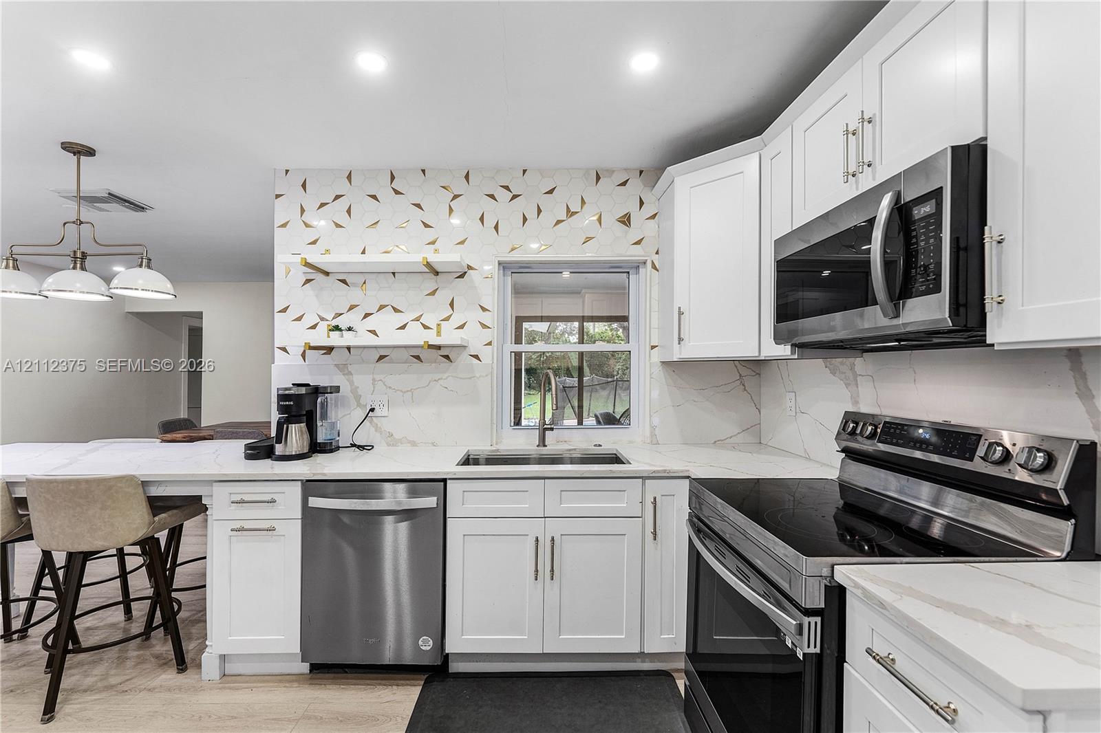 Kitchen featuring white shaker-style cabinetry, stainless steel appliances, patterned tile backsplash, floating shelving with metallic accents, and stone-finish countertops