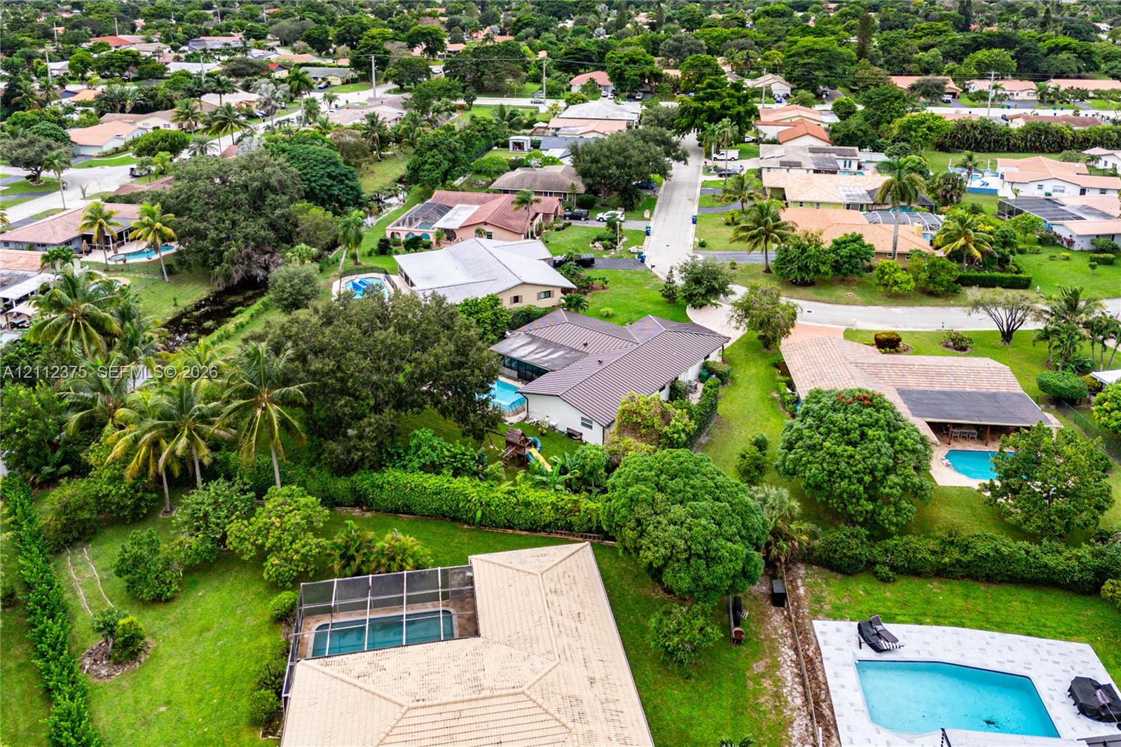 Aerial perspective of a residential property featuring an in-ground swimming pool with a screened enclosure, expansive green space, mature landscaping, and a concrete driveway