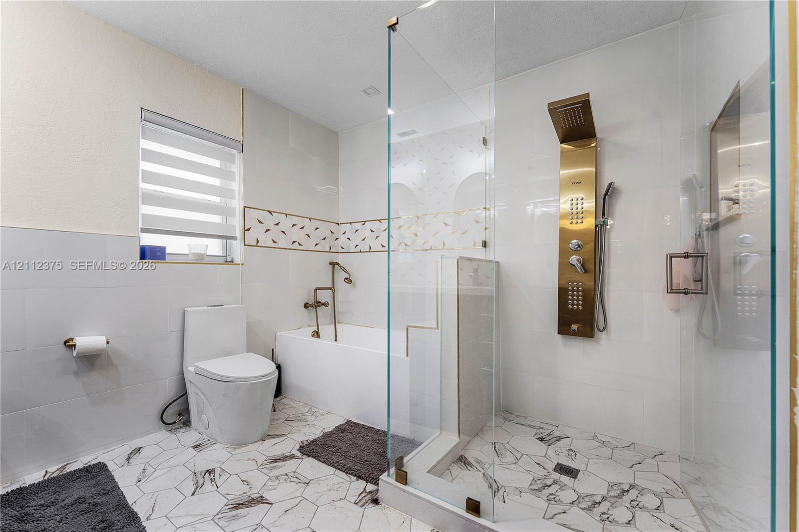 Bathroom featuring a walk-in shower with a gold-tone rainfall shower panel, frameless glass enclosure, a deep soaking tub with a wall-mounted faucet, and hexagonal patterned tile flooring