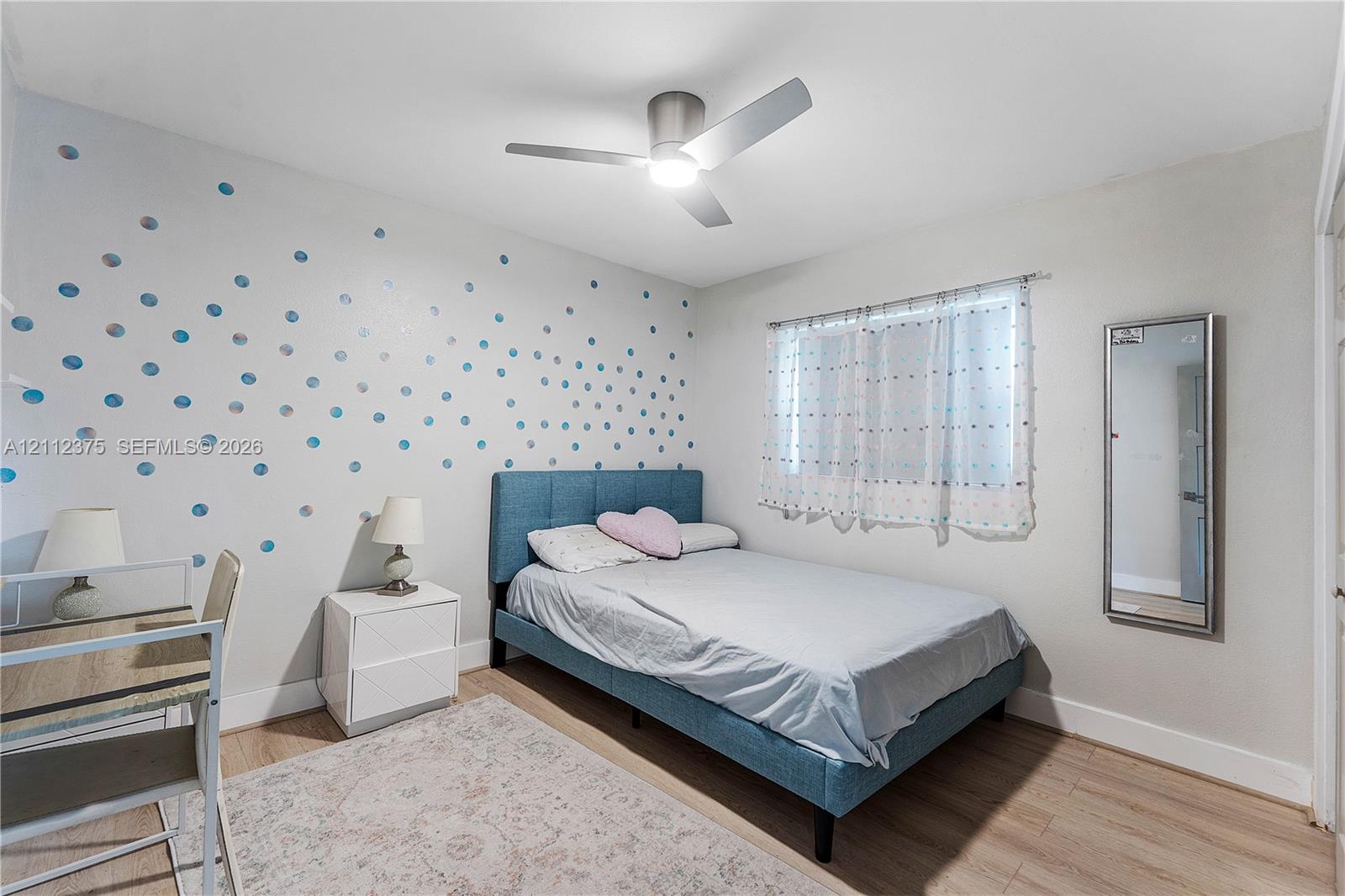 Neutral-toned bedroom featuring wood-finish flooring, a contemporary brushed-metal ceiling fan with integrated lighting, and a wall-mounted mirror