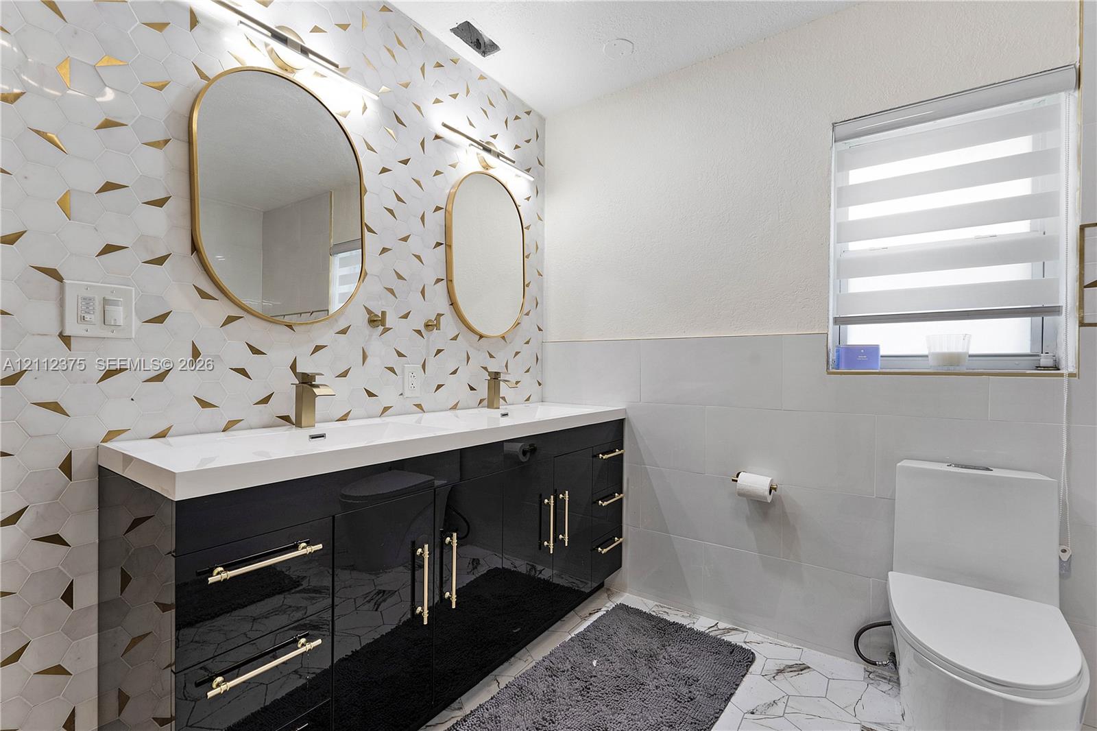 Double vanity featuring a high-gloss black cabinet, gold-tone hardware, rectangular integrated sinks, and quartz-finish countertops, accented by geometric patterned wall tile and dual oval gold-framed mirrors