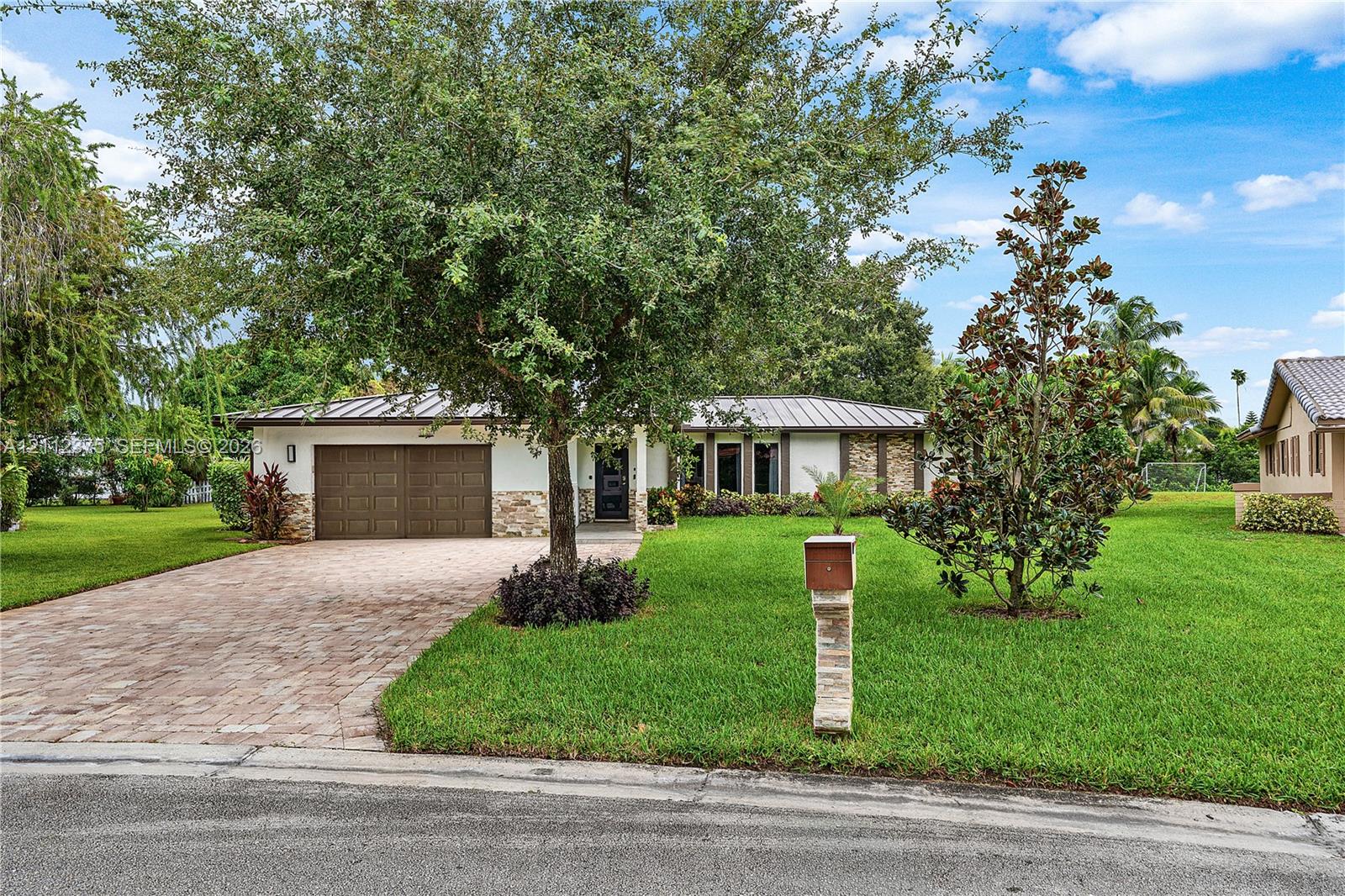 Single-story residence featuring a metal roof, paver driveway, two-car garage, stone veneer accents, and extensive lawn landscaping