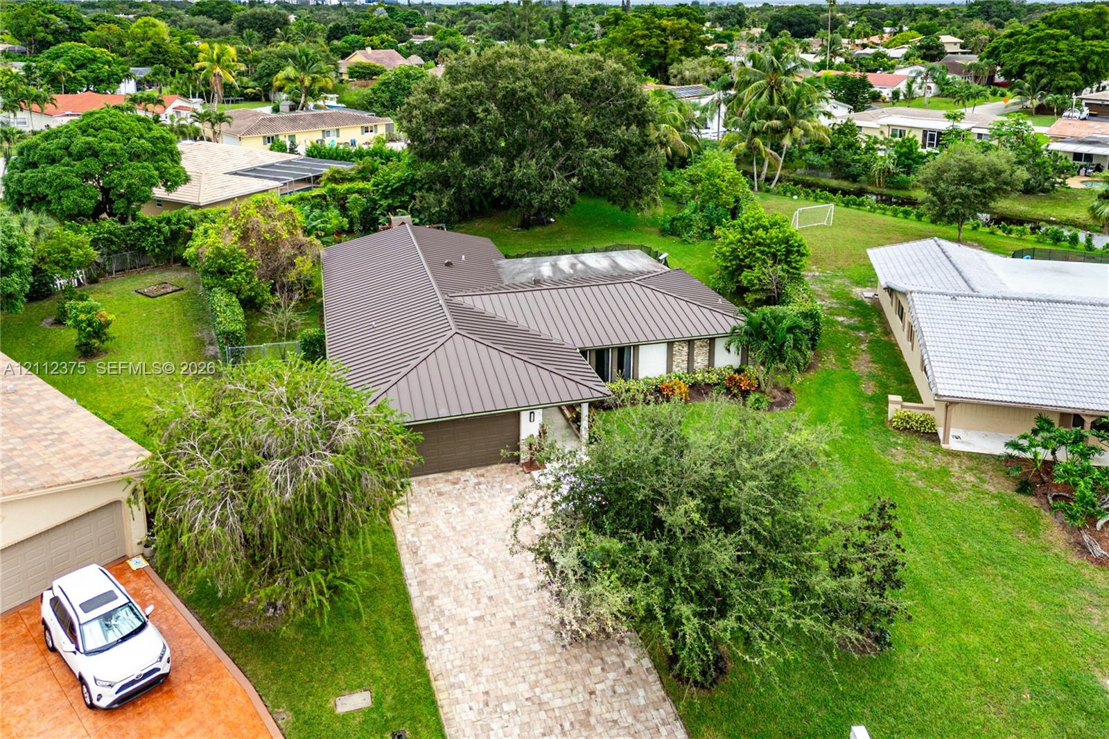 Aerial perspective featuring a metal roof, stone-paved driveway, and expansive lawn with mature landscaping