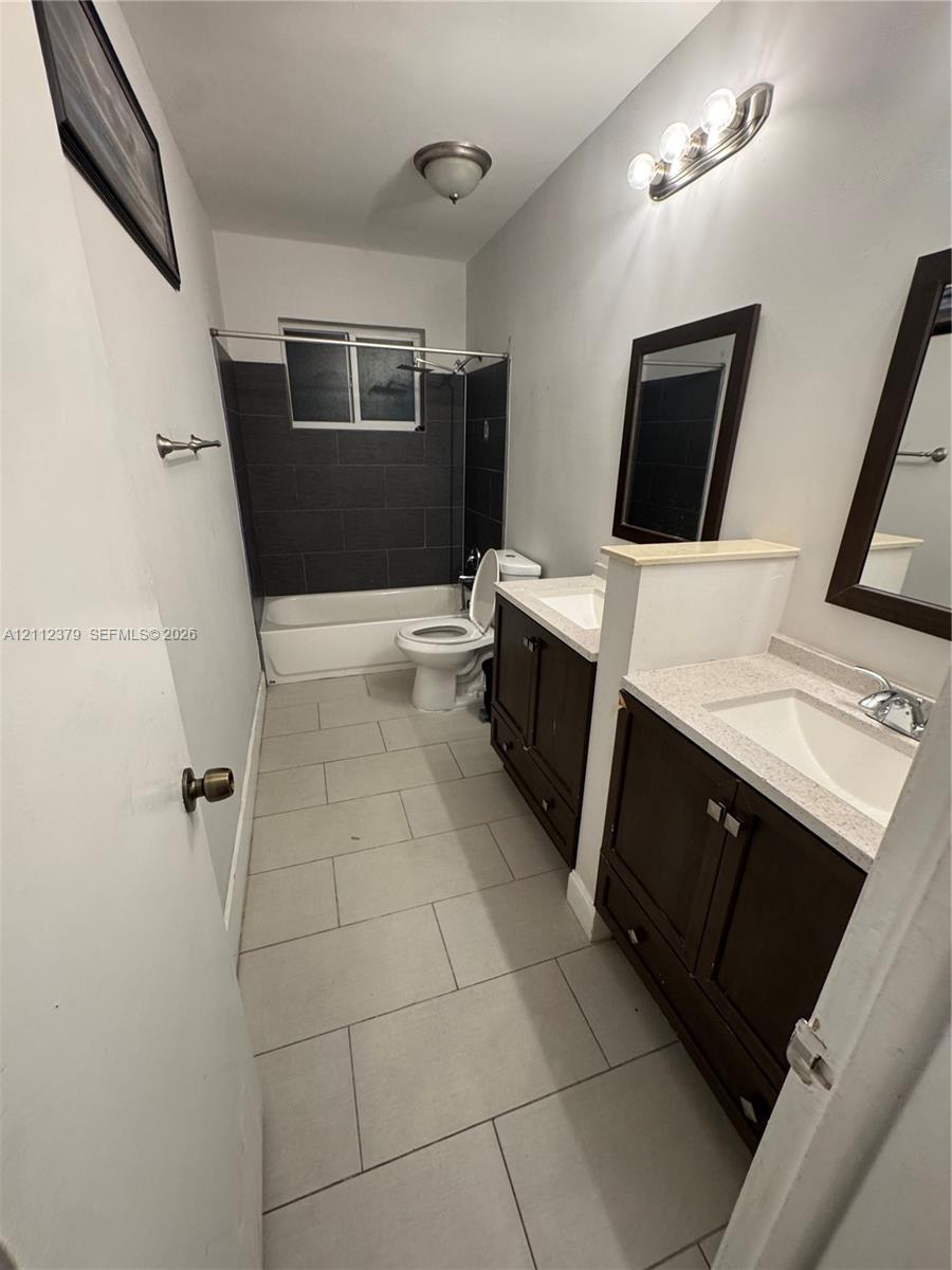 Dual-sink vanity with stone-finish countertops, dark wood cabinetry, and large-format tile flooring, featuring a bathtub with dark tile surround