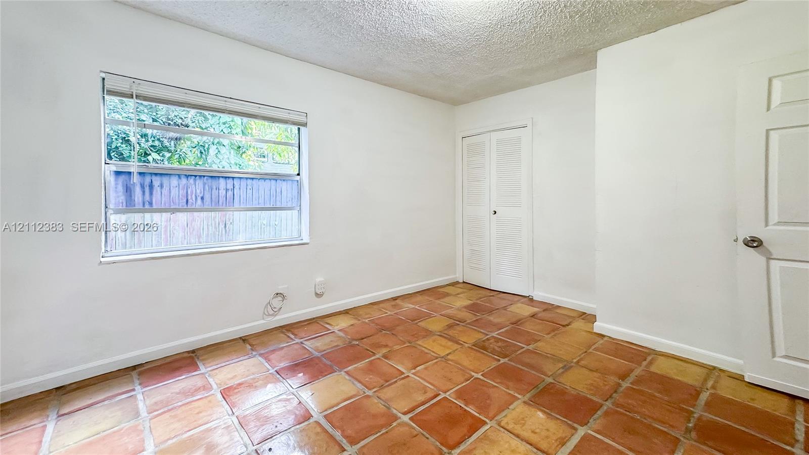 Interior room featuring terracotta tile flooring, white walls, dual louvered closet doors, and a single-hung window with a horizontal blind