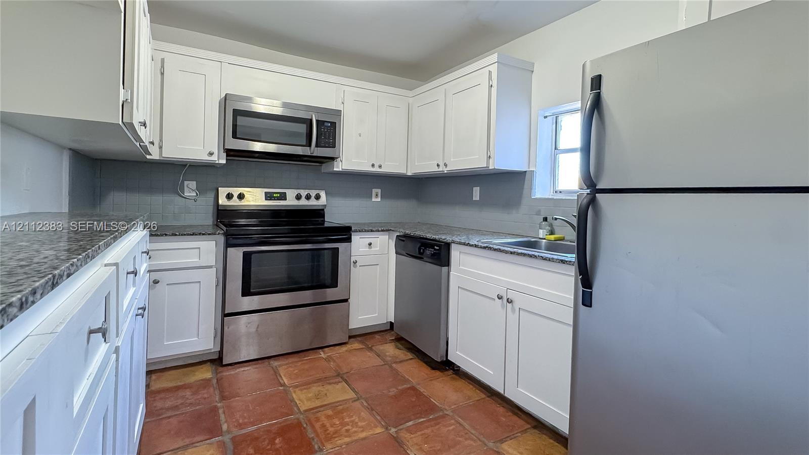 Kitchen featuring white shaker-style cabinetry, speckled stone-finish countertops, stainless steel appliances, and terracotta-style tile flooring
