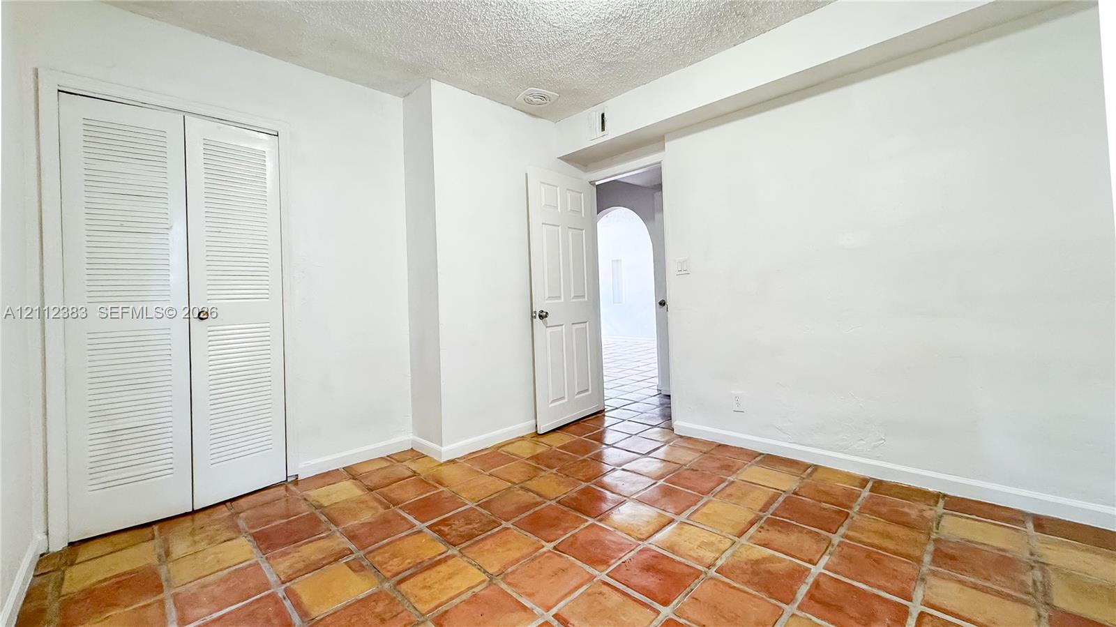 Interior space featuring terracotta-style tile flooring, double louvered closet doors, and a paneled interior door with an arched entryway visible beyond
