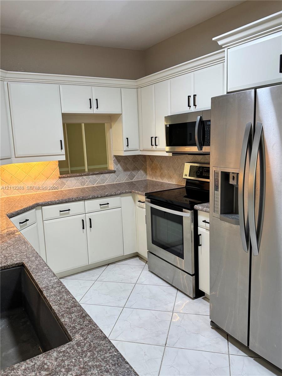 Kitchen featuring white cabinetry with dark hardware, stone-finish countertops, stainless steel appliances, ceramic tile backsplash, and light-toned tile flooring