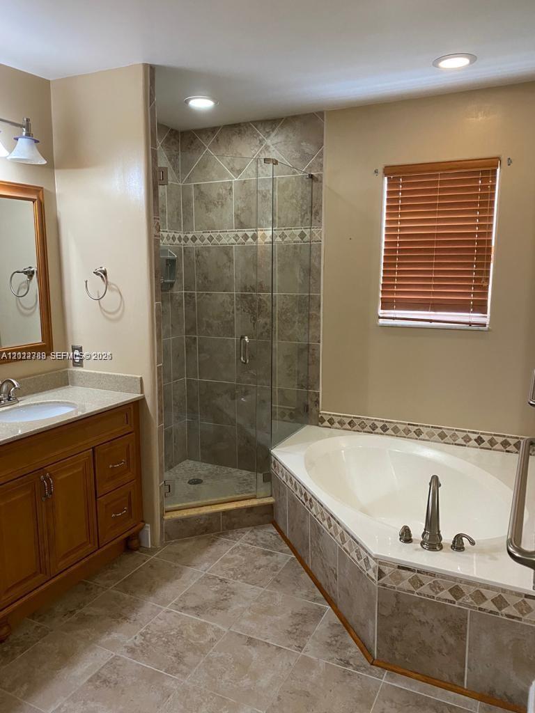 Bathroom featuring a wooden vanity, soaking tub, glass-enclosed shower, stone-finish tile flooring, and decorative tile accents