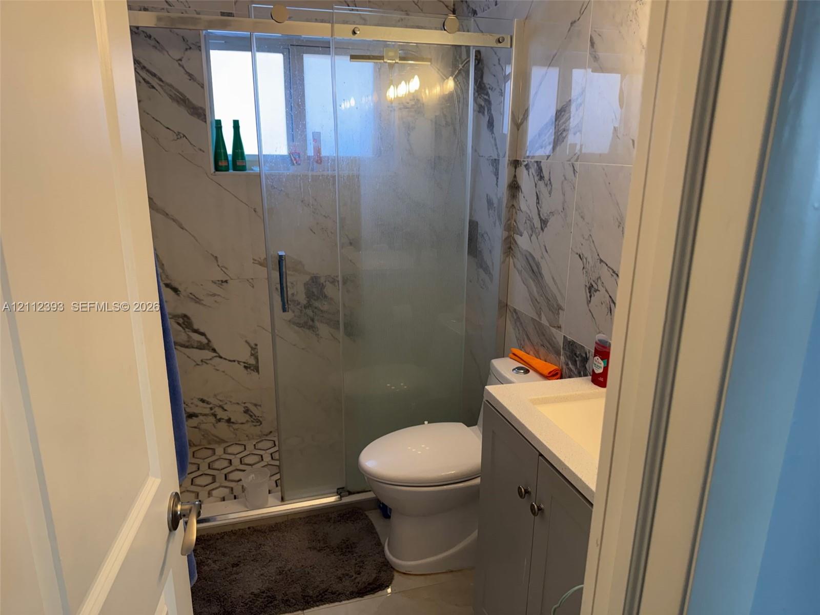 Bathroom featuring a glass-enclosed shower with sliding hardware, large-format marbled tile wall surrounds, decorative hexagon floor tile, and a grey vanity with an integrated countertop sink