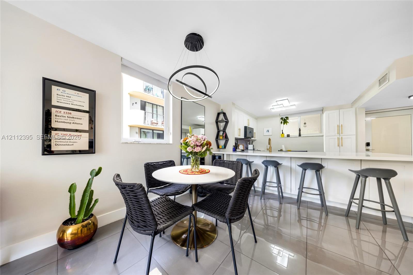 Open-concept kitchen and dining area featuring large-format tile flooring, white cabinetry with gold-tone hardware, a breakfast bar, and a modern tiered ring chandelier