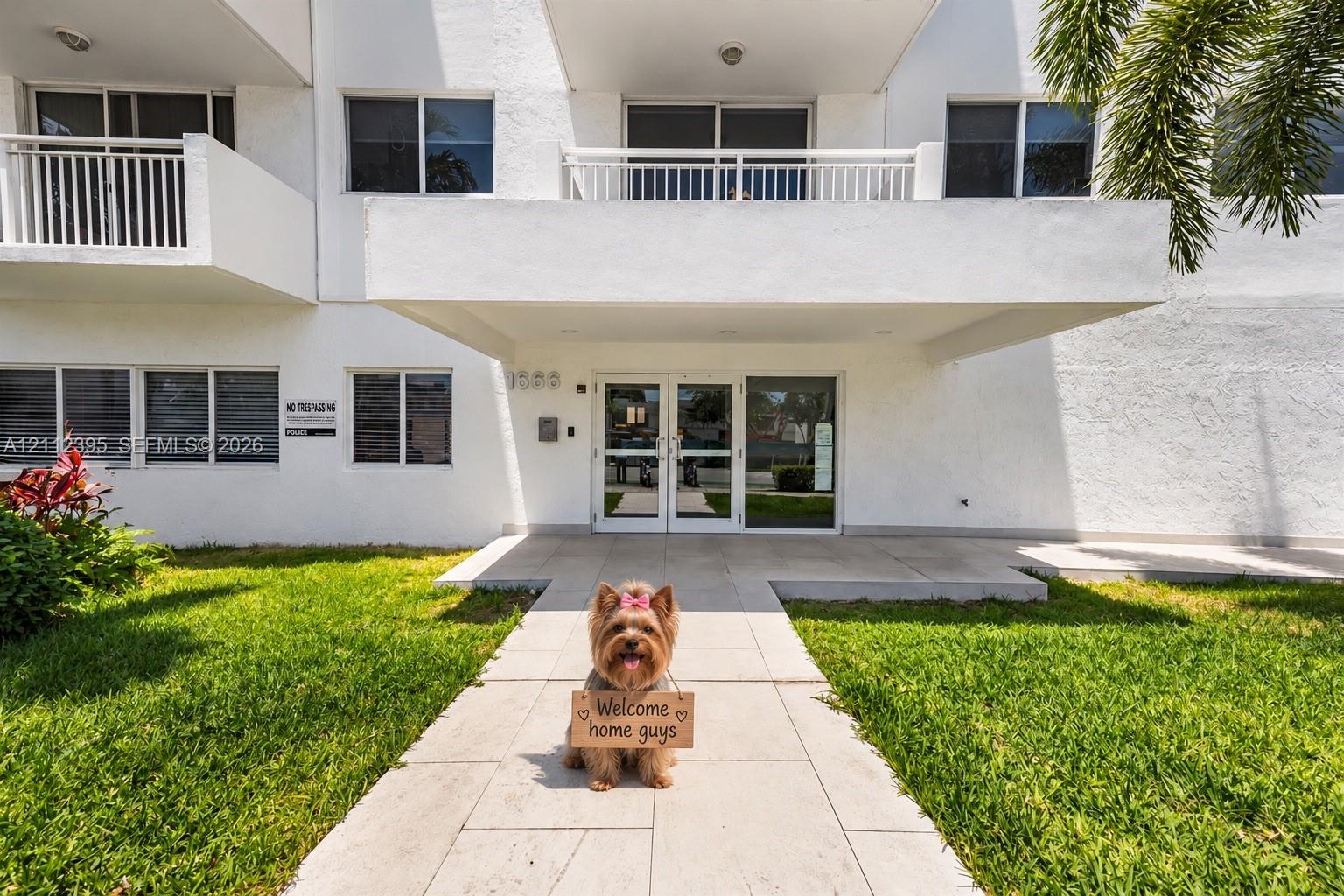 Multi-story residential building exterior featuring a covered entryway, dual glass-panel entrance doors, second-level balconies with white railings, and concrete paver walkways
