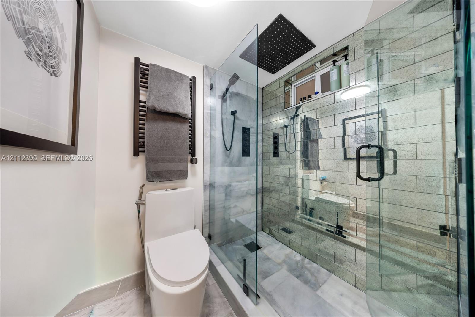 Walk-in shower with stone-tile surround, glass enclosure, and rainfall showerhead, complemented by a matte black heated towel rail and wall-mounted plumbing fixtures