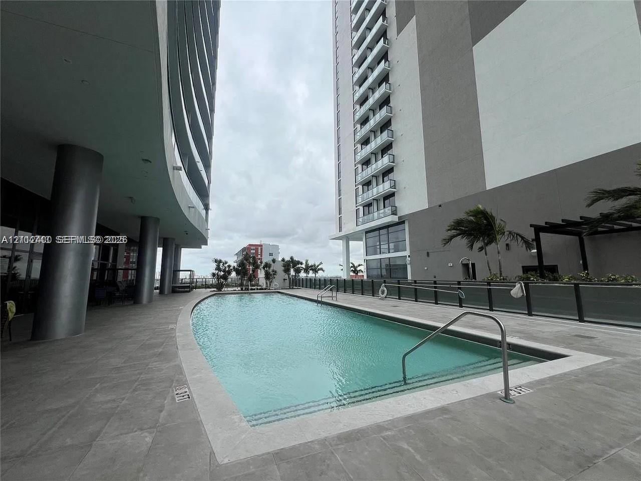 Curved swimming pool featuring a concrete deck, metal handrails, and a glass-panel perimeter fence Curved swimming pool featuring a concrete deck, metal handrails, and a glass-panel perimeter fence