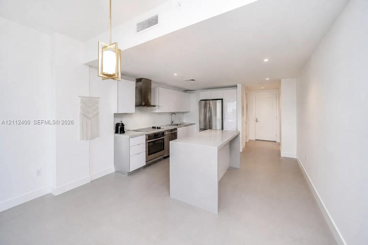 Kitchen featuring flat-panel white cabinetry, a waterfall-edge quartz island, stainless steel appliances, a metallic range hood, and pendant lighting