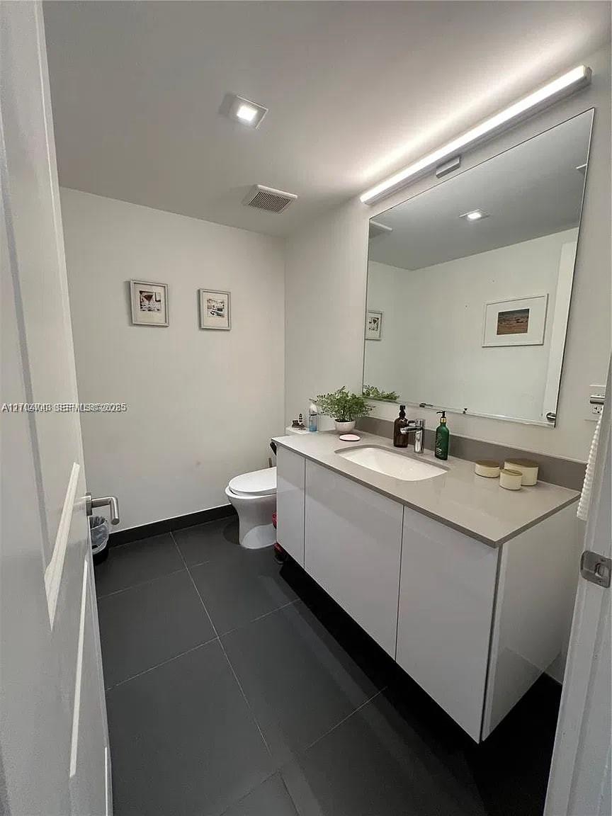 Floating white vanity with solid-surface countertop and integrated sink, large frameless vanity mirror with integrated horizontal light fixture, and dark-toned floor tiling Floating white vanity with solid-surface countertop and integrated sink, large frameless vanity mirror with integrated horizontal light fixture, and dark-toned floor tiling