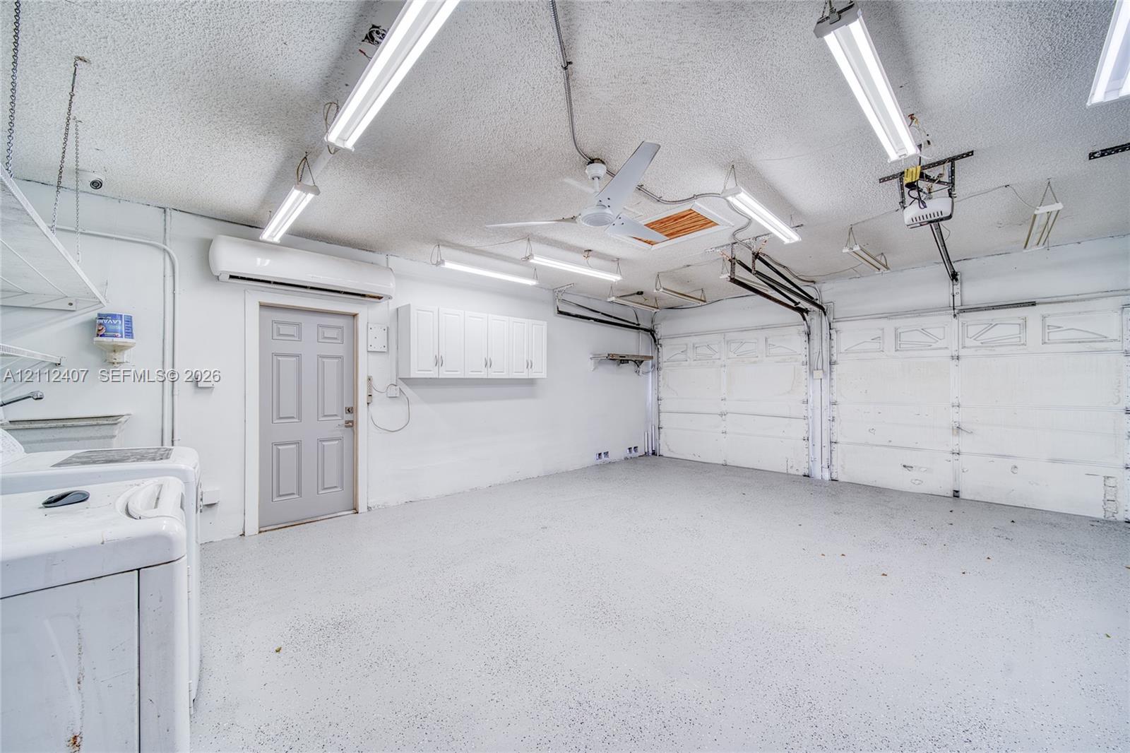 Garage interior featuring epoxy-coated flooring, wall-mounted cabinetry, a ceiling-mounted fan, overhead fluorescent lighting, and a climate-control unit