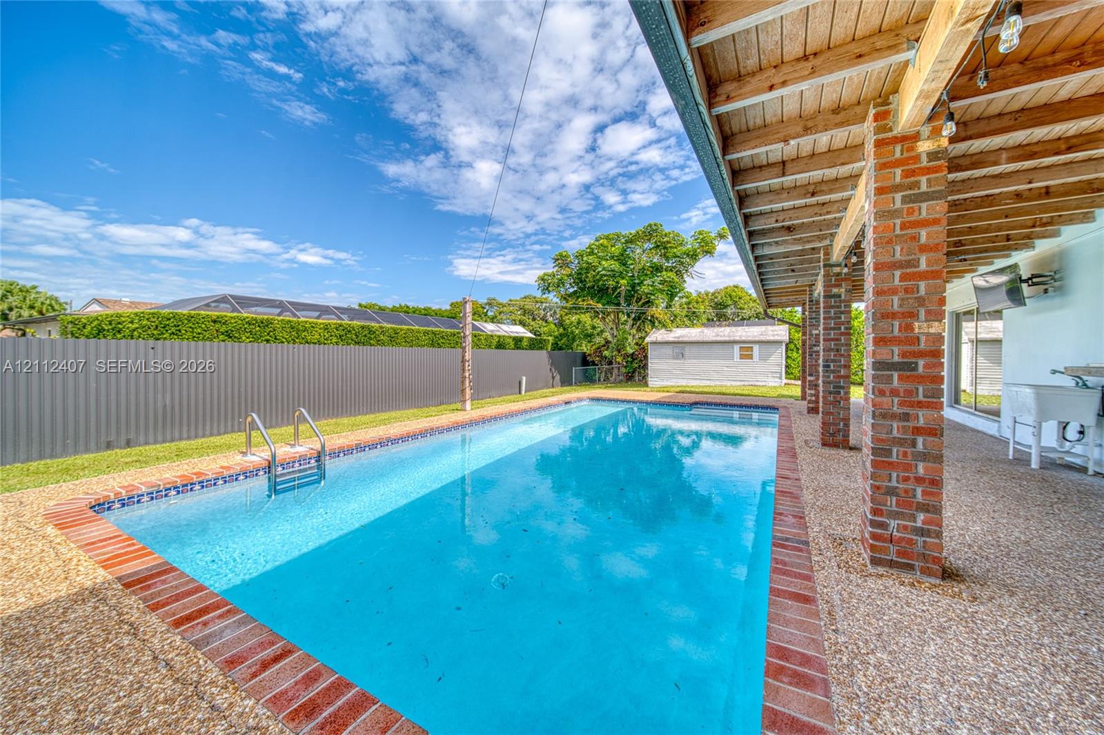 Rectangular in-ground swimming pool featuring a brick surround, covered patio with exposed wood ceiling beams, exposed brick structural pillars, and a metal privacy fence Rectangular in-ground swimming pool featuring a brick surround, covered patio with exposed wood ceiling beams, exposed brick structural pillars, and a metal privacy fence