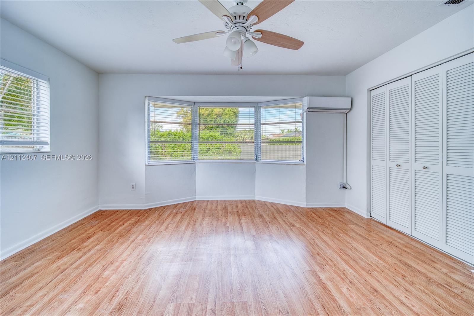 Room featuring wood-finish flooring, a three-panel bay window, wall-mounted ductless climate control unit, ceiling fan with light fixture, and floor-to-ceiling closets with louvered doors