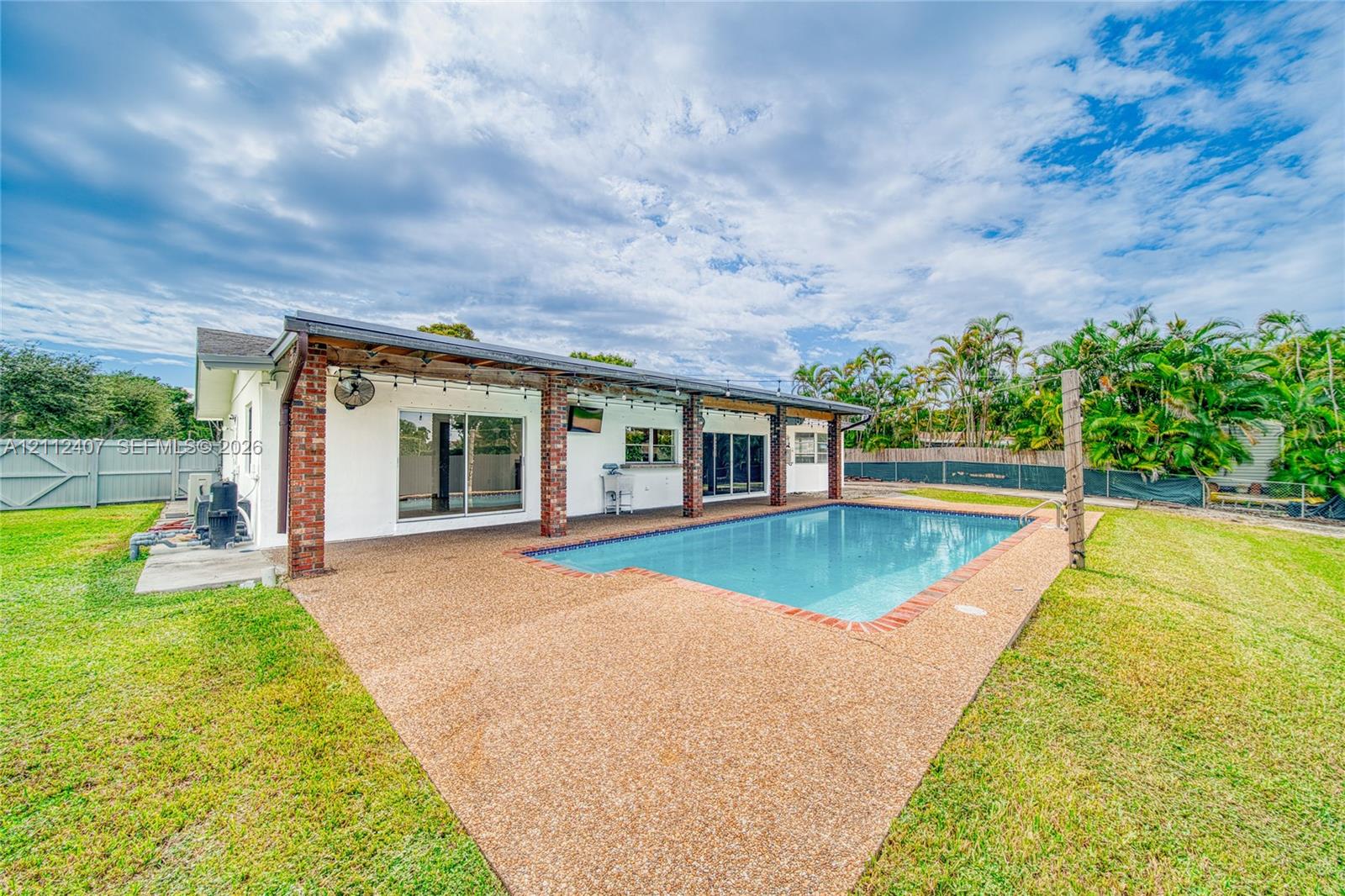 Rear exterior featuring a covered patio with brick support columns, an in-ground pool with brick coping, expansive lawn, and fenced perimeter Rear exterior featuring a covered patio with brick support columns, an in-ground pool with brick coping, expansive lawn, and fenced perimeter
