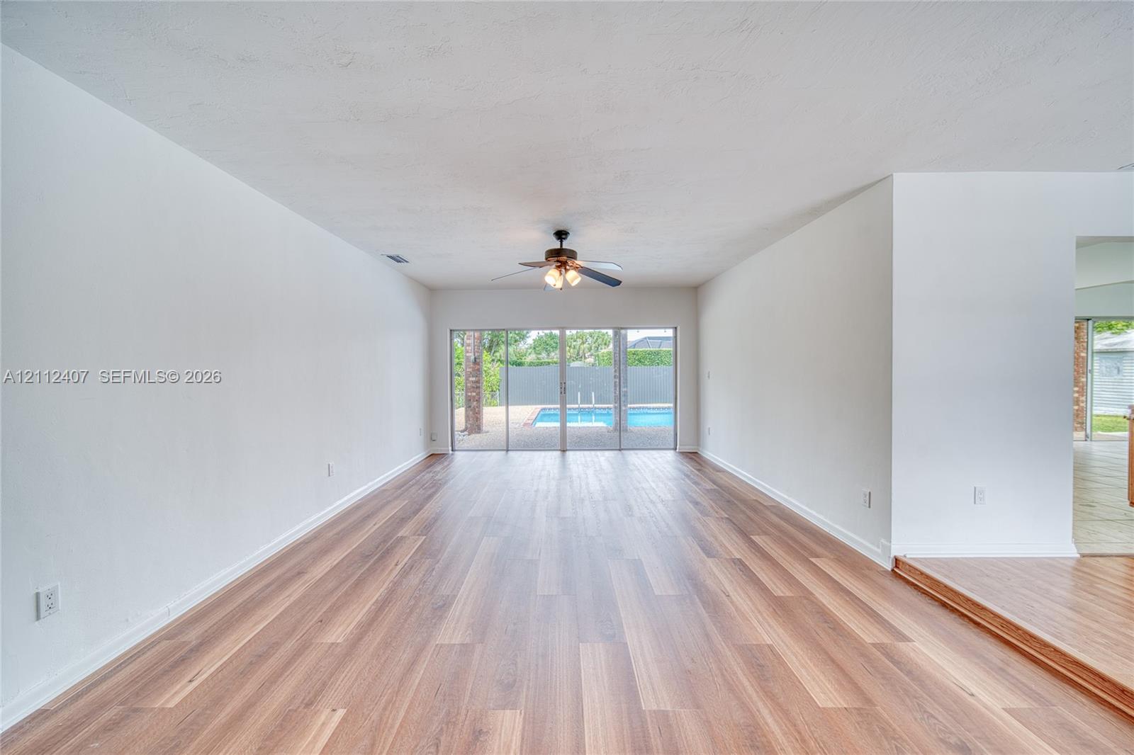 Interior living space featuring wood-finish flooring, a ceiling fan with integrated lighting, and glass sliding doors revealing a swimming pool Interior living space featuring wood-finish flooring, a ceiling fan with integrated lighting, and glass sliding doors revealing a swimming pool