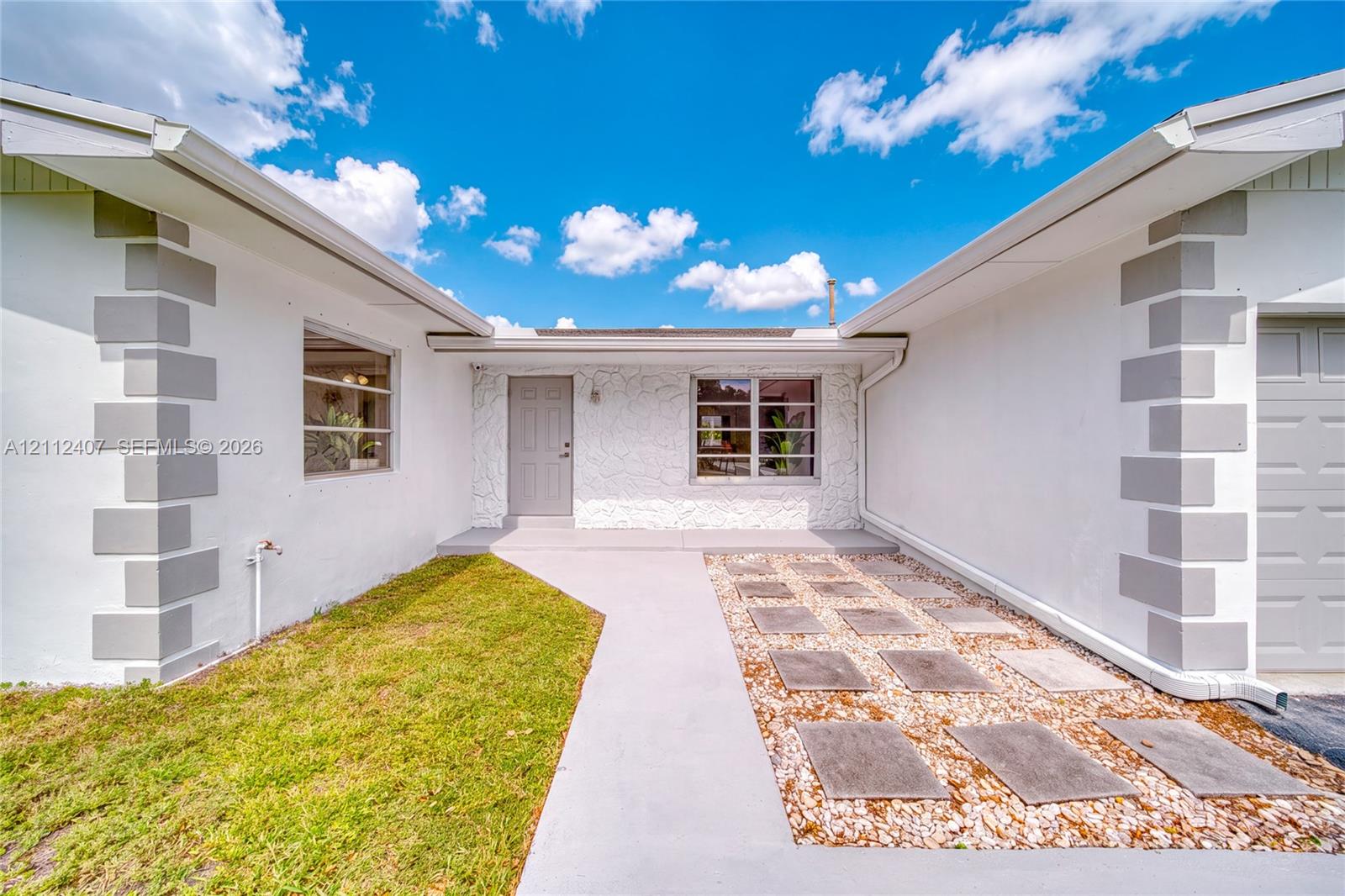 Exterior featuring white stucco finish with grey decorative quoins, textured stone masonry accents, a concrete walkway, and a geometric hardscape design Exterior featuring white stucco finish with grey decorative quoins, textured stone masonry accents, a concrete walkway, and a geometric hardscape design