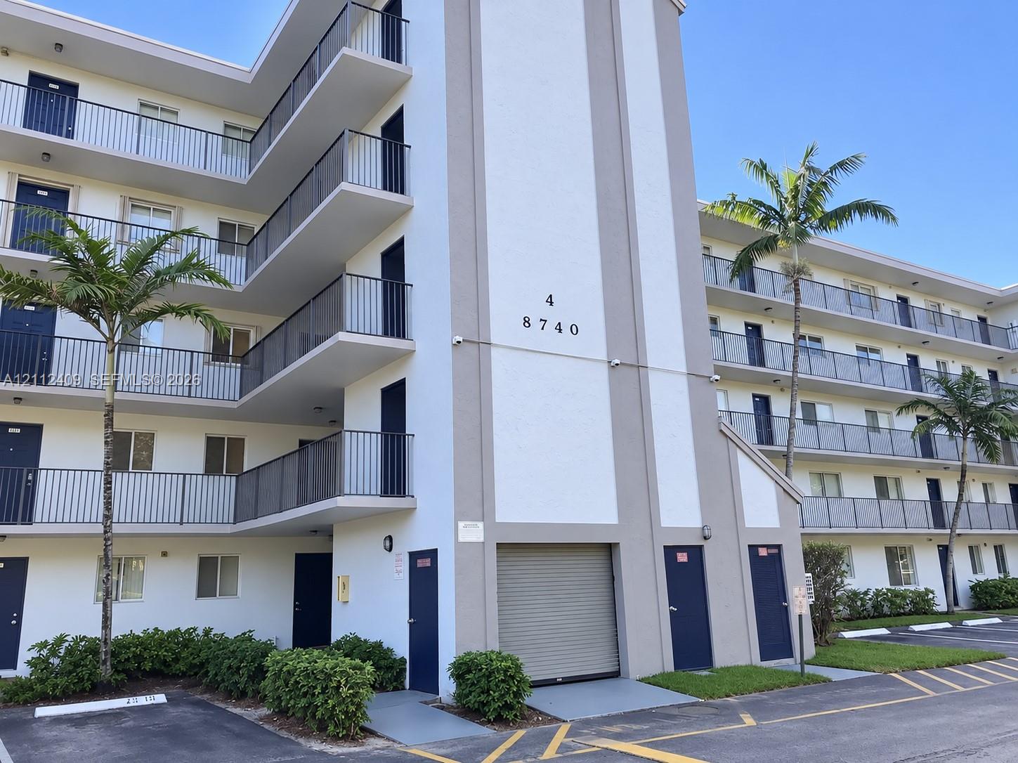 Multi-story building exterior featuring stacked private balconies, metal railings, a ground-level roll-up security door, and exterior landscaping Multi-story building exterior featuring stacked private balconies, metal railings, a ground-level roll-up security door, and exterior landscaping
