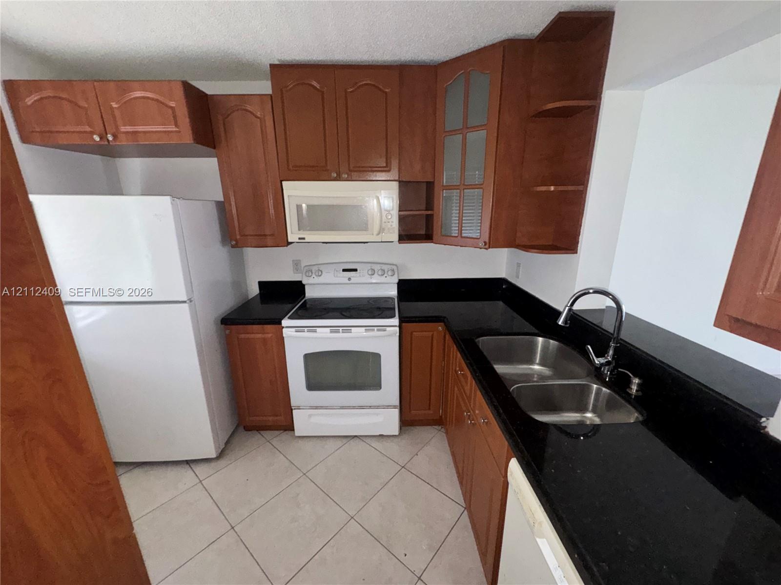 Kitchen featuring wood-finish cabinetry, solid surface countertops, a stainless steel double-basin sink with a high-arch faucet, white appliances, and tiled flooring