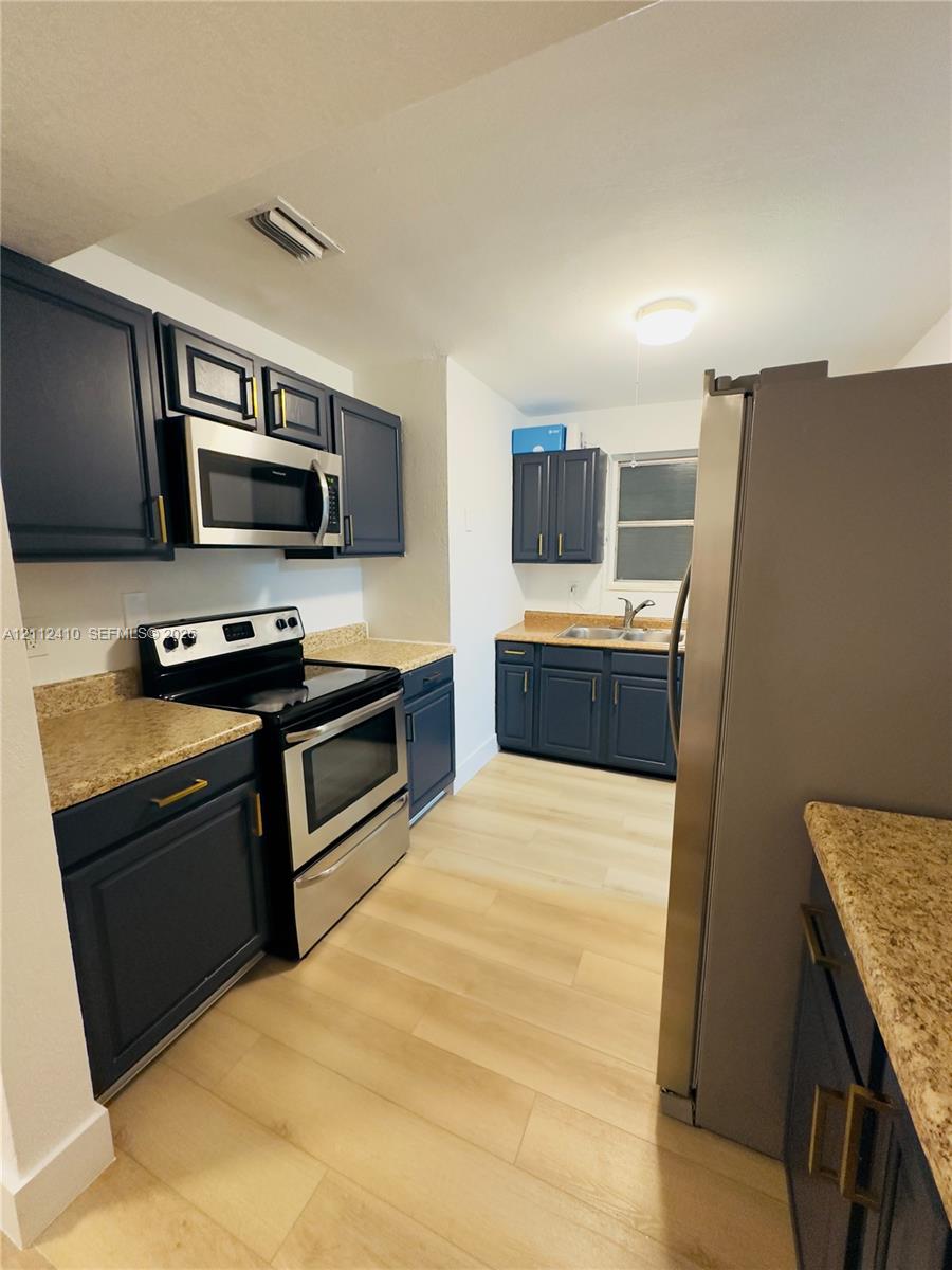 Kitchen featuring navy blue shaker-style cabinetry, gold-tone hardware, stone-finish countertops, light wood-finish flooring, and stainless steel appliances