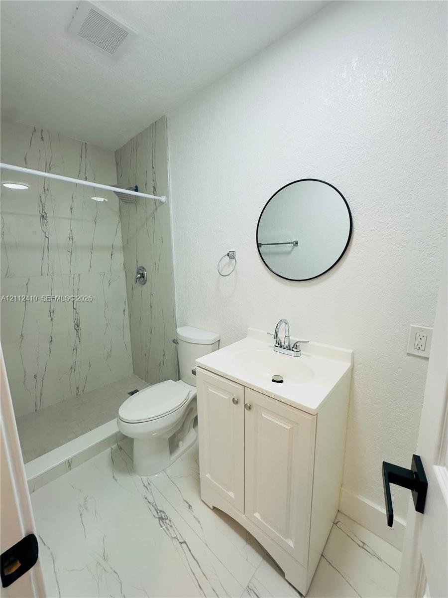 Bathroom featuring a walk-in shower with stone-finish tile surround, white vanity cabinet with a drop-in sink, circular wall-mounted mirror, and large-format tile flooring