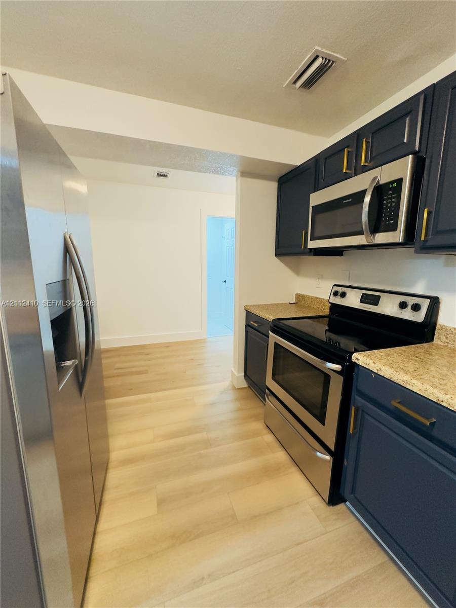 Kitchen featuring navy blue cabinetry with metallic hardware, stone-finish countertops, stainless steel appliances, and light-toned wood-finish flooring