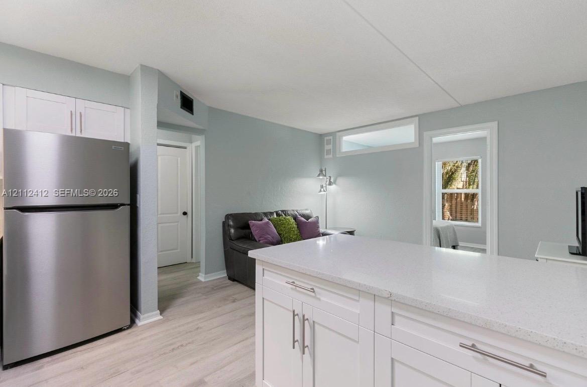 Kitchen area featuring white cabinetry, a quartz countertop island, stainless steel refrigerator, and wood-finish flooring Kitchen area featuring white cabinetry, a quartz countertop island, stainless steel refrigerator, and wood-finish flooring