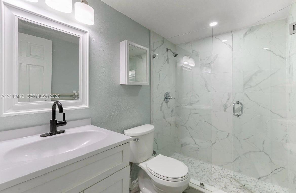 Bathroom featuring a white vanity cabinet, matte black faucet, wall-mounted medicine cabinet, and a walk-in shower with floor-to-ceiling stone-patterned tile and a frameless glass door Bathroom featuring a white vanity cabinet, matte black faucet, wall-mounted medicine cabinet, and a walk-in shower with floor-to-ceiling stone-patterned tile and a frameless glass door