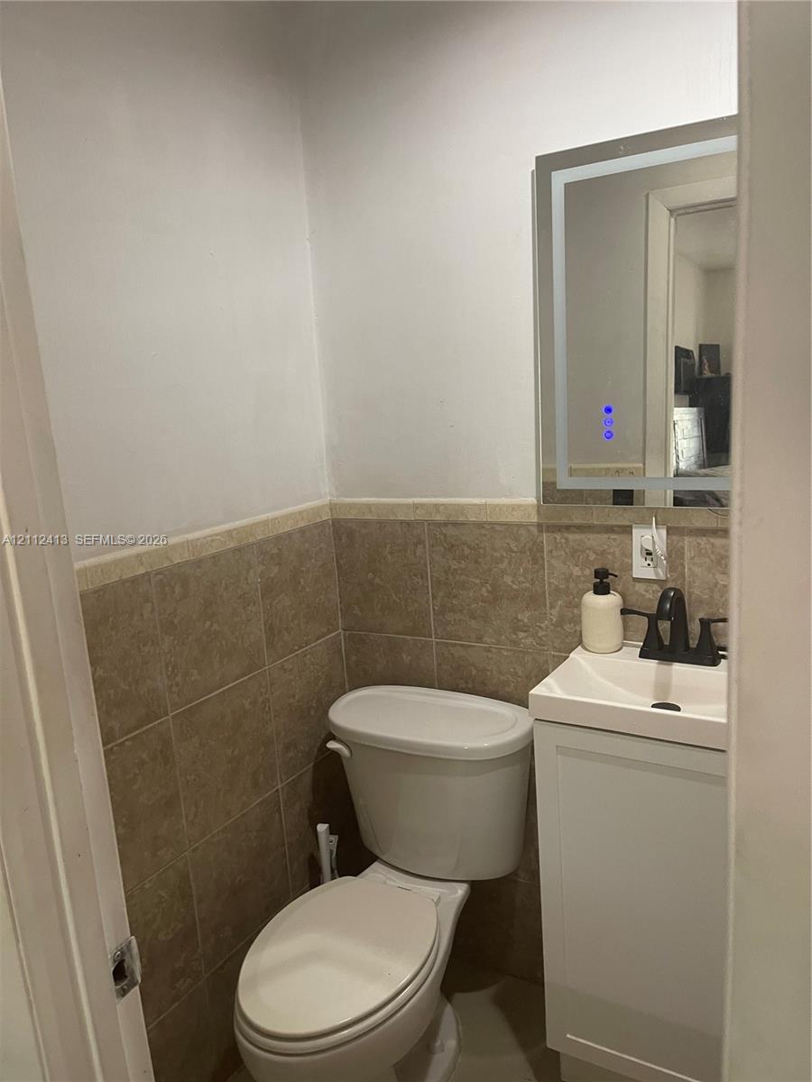 Wall-mounted LED backlit mirror, pedestal-style vanity with a rectangular sink, matte black faucet, and tiled wainscoting
