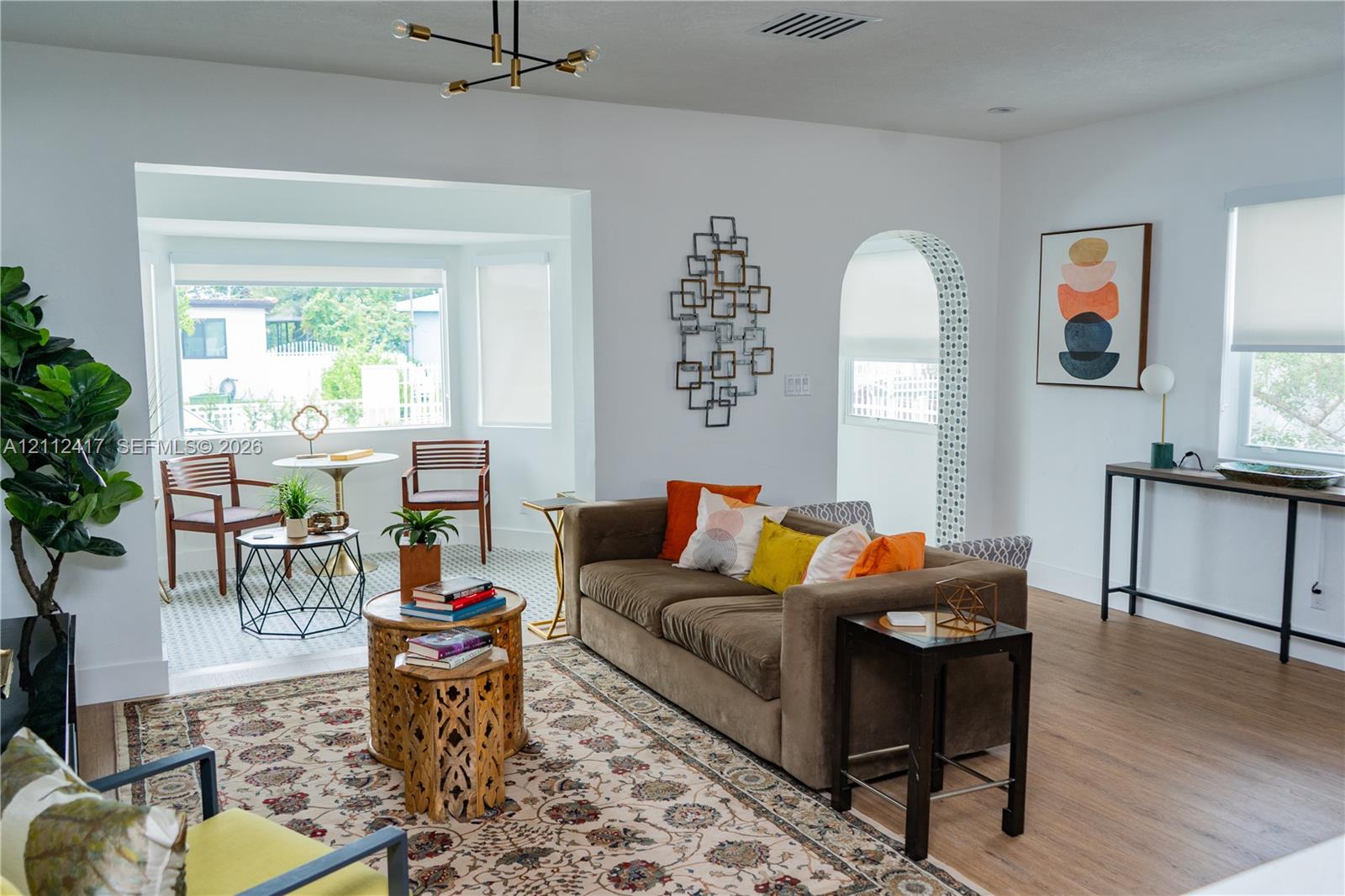 Living area featuring wood-finish flooring, a bay window with natural light, a distinctive arched doorway with patterned tile surround, and a modern multi-arm ceiling light fixture