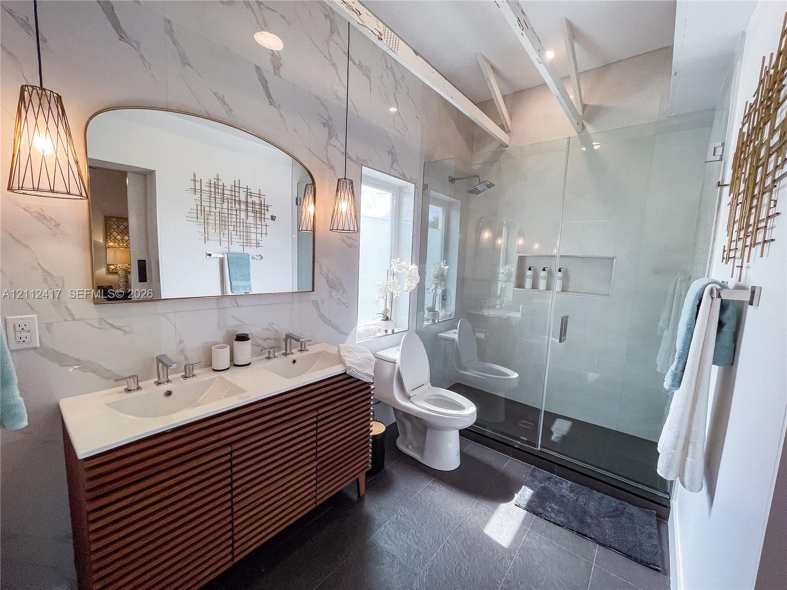 Bathroom featuring a dual-sink vanity with slatted wood cabinet doors, large format stone-finish wall tile, a walk-in glass-enclosed shower with a built-in niche, and dual pendant light fixtures
