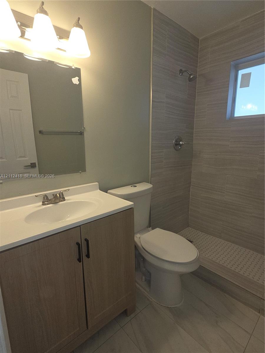 Wood-finish vanity with a composite countertop, wall-mounted mirror, light fixture, walk-in shower with horizontal tile surround, and tile flooring Wood-finish vanity with a composite countertop, wall-mounted mirror, light fixture, walk-in shower with horizontal tile surround, and tile flooring