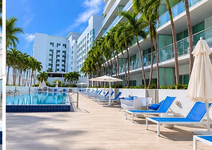 Swimming pool featuring a raised tiled edge, extensive paved pool deck, glass balcony railings, and mature palm trees