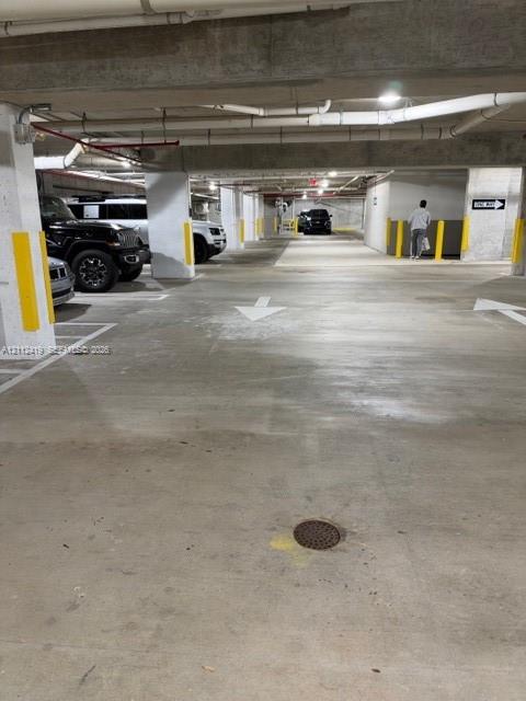 Covered parking garage with concrete flooring, structural pillars featuring yellow protective markers, and painted directional signage