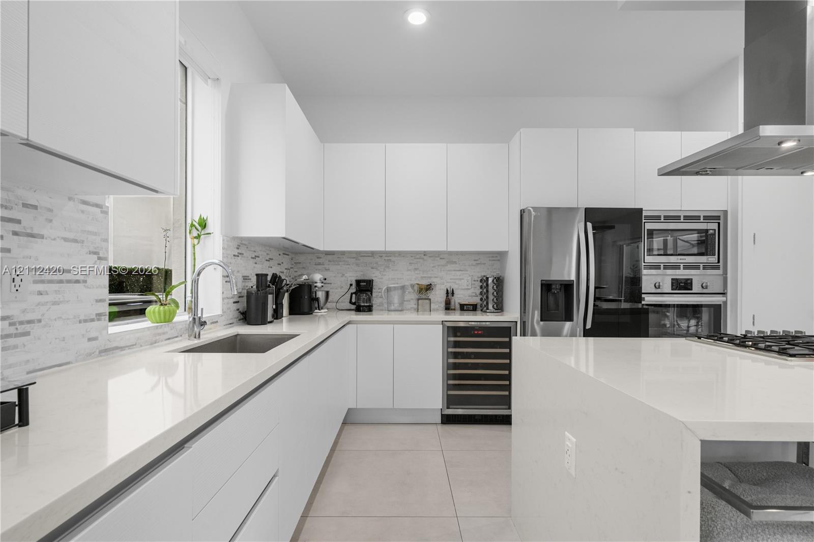 Modern kitchen featuring white flat-panel cabinetry, quartz countertops, a tiled backsplash, stainless steel appliances including a built-in wine refrigerator, and a waterfall-edge kitchen island