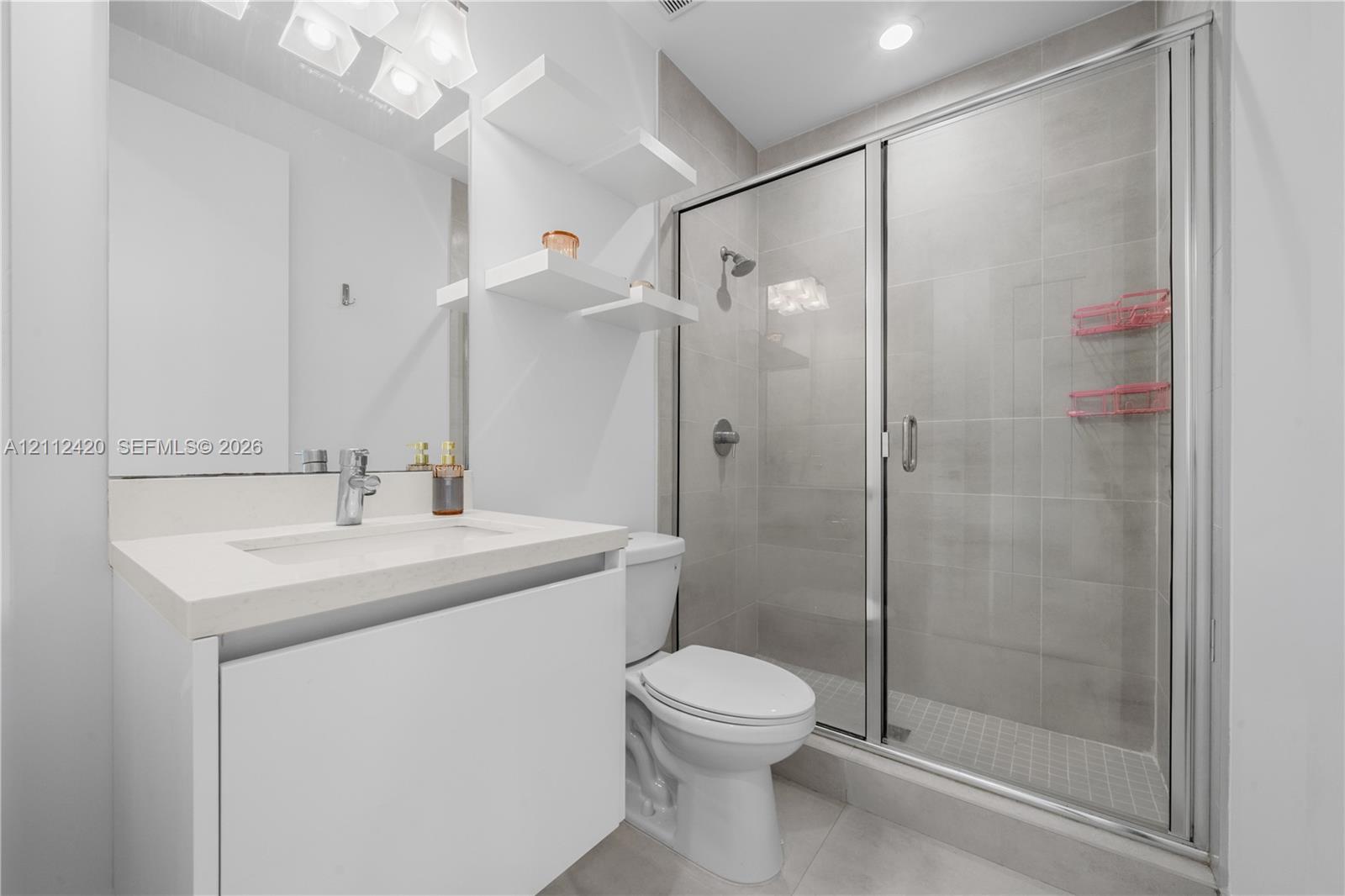 Bathroom featuring a white vanity with a rectangular sink, quartz countertop, built-in wall shelving, and a tiled shower enclosure with a glass door