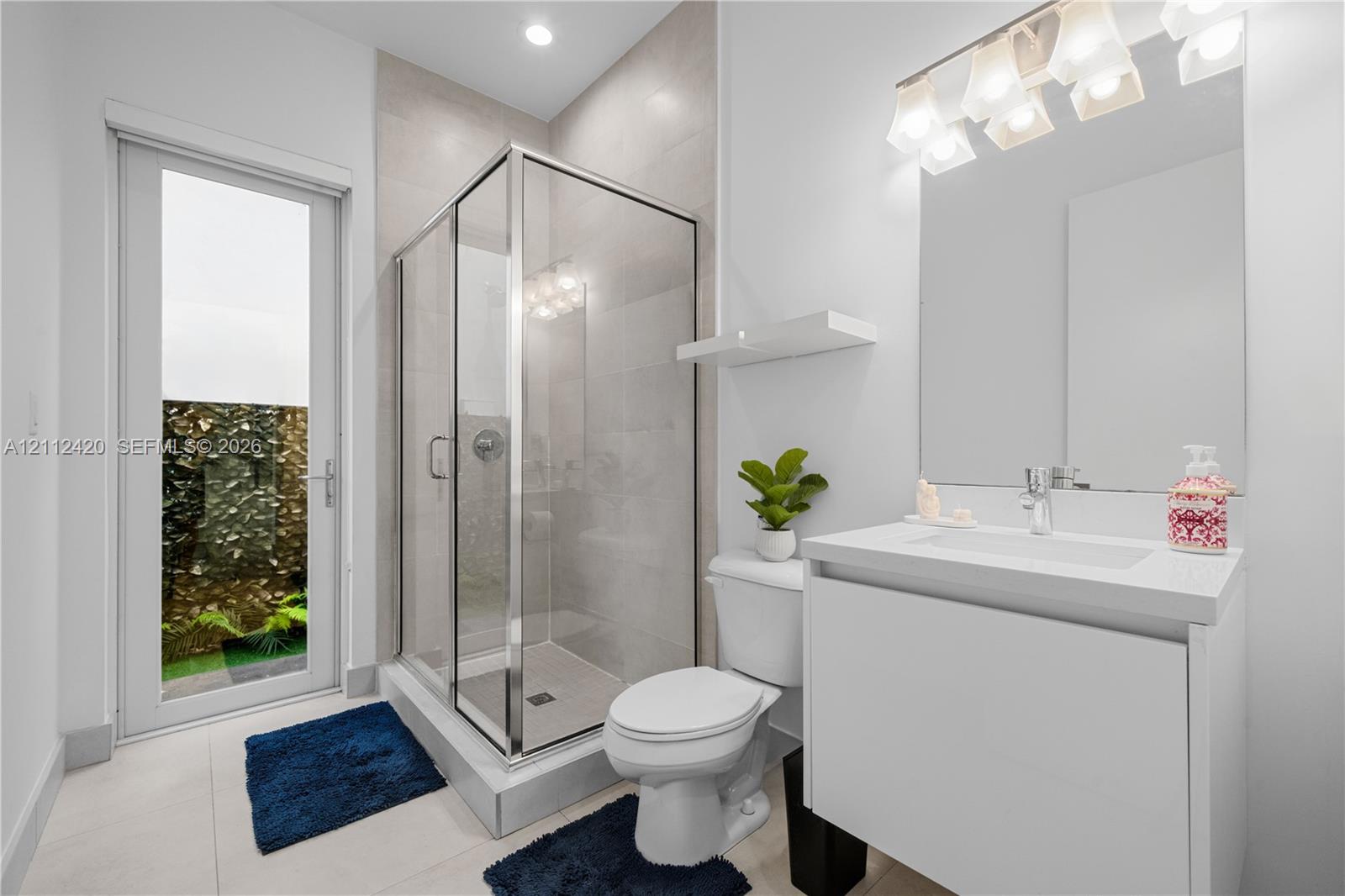 Full-height tiled walk-in shower with glass enclosure, white floating vanity with integrated rectangular sink, stone-finish countertop, and glass-paneled exterior door