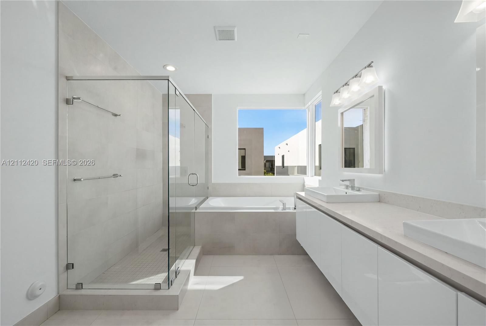 Bathroom featuring a glass-enclosed walk-in shower with tiled surround, a built-in soaking tub, a long dual-sink vanity with light-tone countertops, and high-gloss white cabinetry