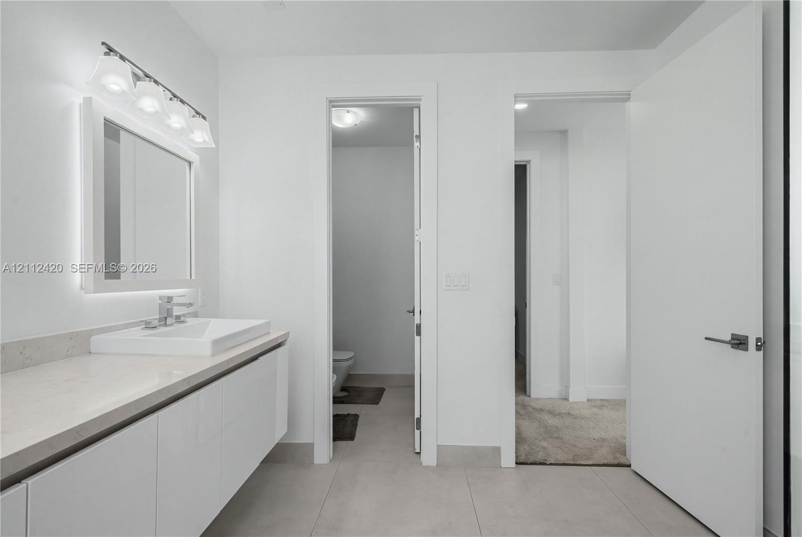 Modern floating vanity with a rectangular sink, light-toned countertop, four-bulb vanity light, and large format floor tile