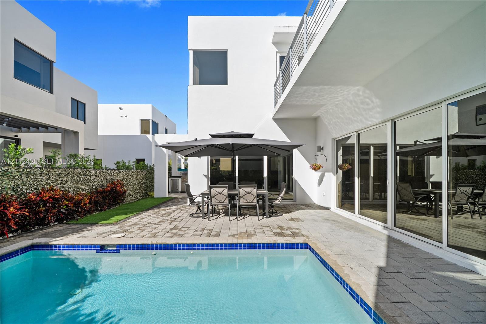Modern stucco exterior with expansive sliding glass doors, paver patio, in-ground pool with blue tile border, and privacy hedge